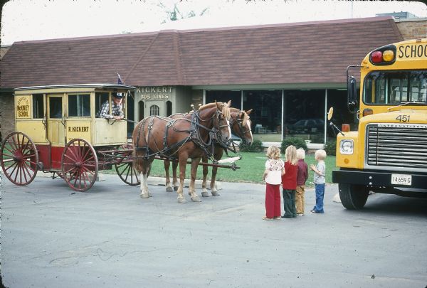 View towards a group of young school children standing in front of a man in a horse-drawn Kickert carriage on a parking area. Signs painted on the side of the carriage read: "Service to McKinley School" and "R. Kickert." On the right is a yellow school bus, and in the background is a building with a sign that reads "Kickert Bus Lines."