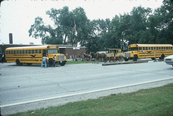 View across road towards Kickert school buses at a school. Children are waiting in a line on the sidewalk. Some of the children are in a horse-drawn carriage that has painted signs on the side that read: "R. Kickert" and "Service to McKinley School."
