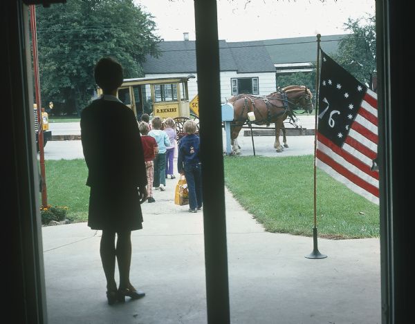 View through school building doorway towards children walking towards Kickert school buses and a horse-drawn carriage. A Bennington flag is visible on a temporary flagpole. A woman is standing near the entrance to the school watching the children.