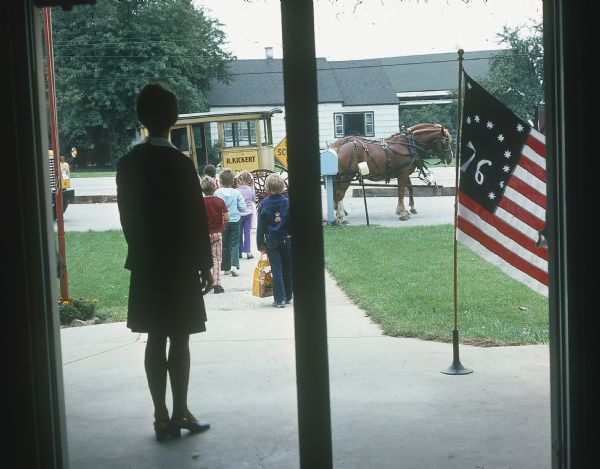 View through school building doorway towards children walking towards Kickert school buses and a horse-drawn carriage. A Bennington flag is visible on a temporary flagpole. A woman is standing near the entrance to the school watching the children. 