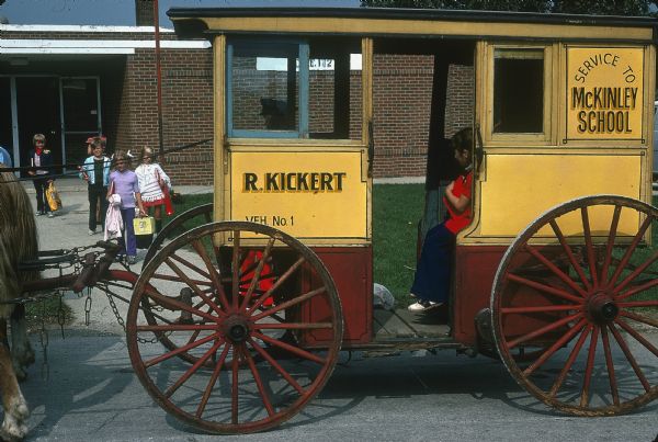 Left side view of Kickert horse-drawn carriage. Signs painted on the side of the carriage read: "Service to McKinley School" and "R. Kickert." One child is sitting in the carriage, and behind the carriage a group of children are walking on the sidewalk in front of a school building.