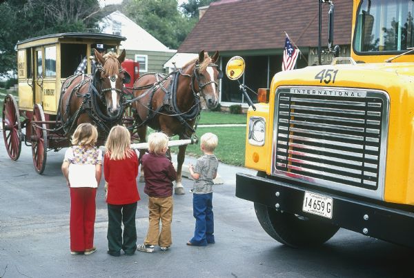 Children standing in a parking lot in front of a horse-drawn carriage. The carriage has painted signs on the side that read: "R. Kickert" and "Service to McKinley School." There is a yellow school bus on the right, and in the background is a building with a sign that reads: "Kickert Bus Lines."