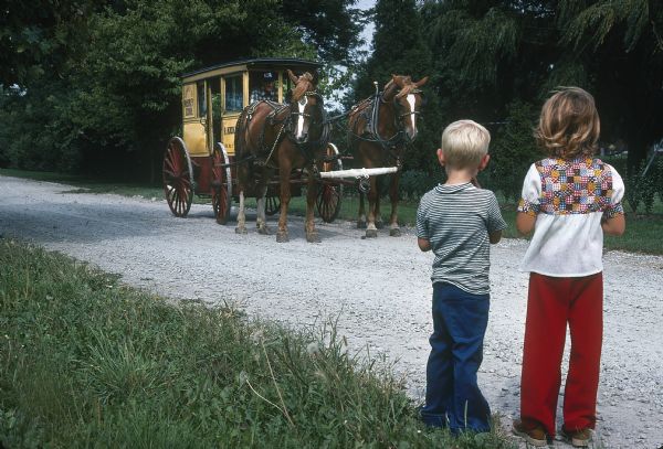 Two young children are standing on the side of a gravel road looking towards a man driving a horse-drawn Kickert carriage.
