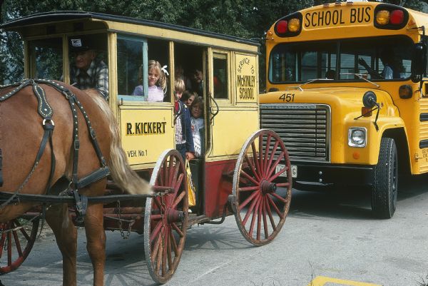 Children are inside a horse-drawn Kickert carriage driven by a man wearing a Kickert hat. Behind the carriage is a yellow school bus.