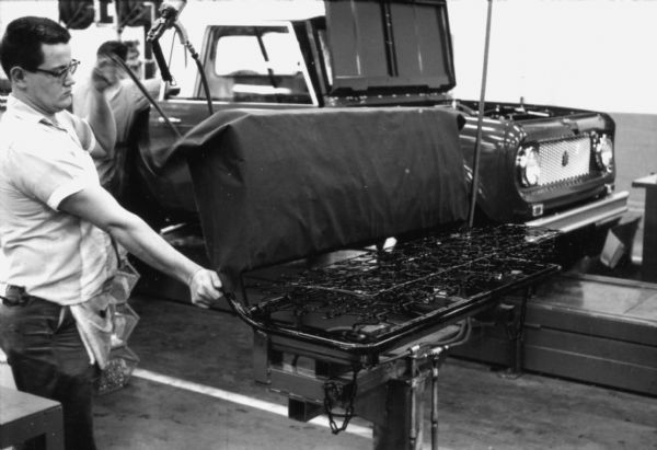 A Fort Wayne employee is in the foreground covering a bench style seat for installation. In the background is an International Scout body, with hood open, moving along an assembly line. Assembly line image from International Harvester's Fort Wayne plant, subject of a 1961 article in <i>International Harvester Today</i> entitled: "The Scout."