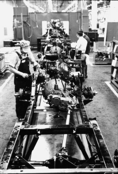 Man working on assembly line with International Scout chassis on the Fort Wayne production line. Two other men are standing in the background on the right. Assembly line image from International Harvester's Fort Wayne plant, subject of a 1961 article in <i>International Harvester Today</i> entitled: "The Scout."