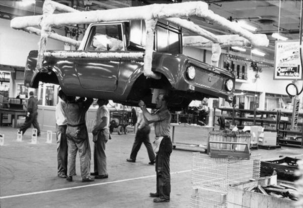Four International Harvester employees standing under an elevated Scout body making final adjustments as they prepare to mount the body on its chassis. Assembly line image from International Harvester's Fort Wayne plant, subject of a 1961 article in <i>International Harvester Today</i> entitled: "The Scout."