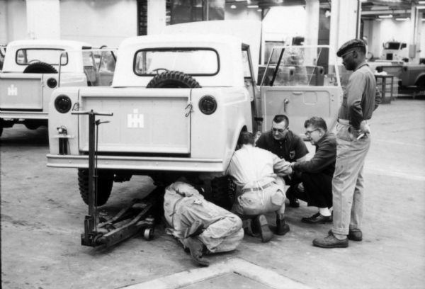 Rear view of an International Scout, with hood up and doors open, suspended on a jack as it nears its complete assembly. There is a spare tire inside the truck bed. One man is lying under the truck bed near the jack. Other men crouch and stand on the right side of the truck. Assembly line image from International Harvester's Fort Wayne plant, subject of a 1961 article in <i>International Harvester Today</i> entitled: "The Scout."