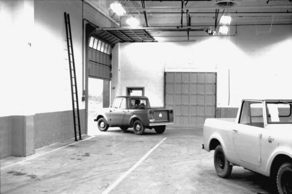 View across factory floor towards an International Scout being driven out of an open garage door of the Fort Wayne factory following its construction. Another Scout is parked in the foreground on the right. Assembly line image from International Harvester's Fort Wayne plant, subject of a 1961 article in <i>International Harvester Today</i> entitled: "The Scout."