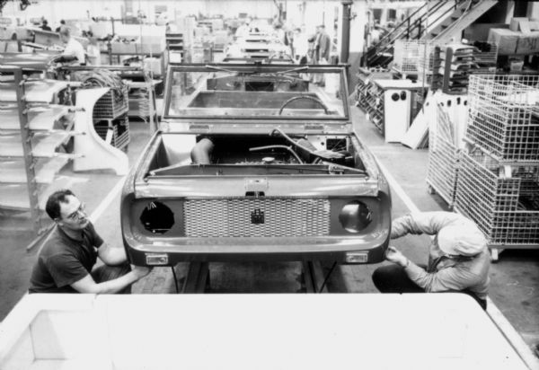 Slightly elevated view looking down center of assembly line towards Fort Wayne employees making adjustments to the front headlights of International Scout body. Assembly line image from International Harvester's Fort Wayne plant, subject of a 1961 article in <i>International Harvester Today</i> entitled: "The Scout."