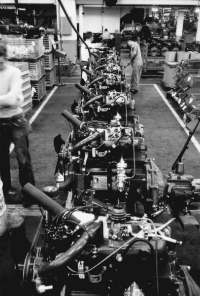 View down row of Scout engines in preparation for installment, Fort Wayne production line. Three men are working near the engines. Assembly line image from International Harvester's Fort Wayne plant, subject of a 1961 article in <i>International Harvester Today</i> entitled: "The Scout."