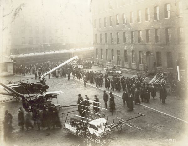 Elevated view looking down at men looking at agricultural equipment outdoors in a paved area near brick buildings. There are railroad cars in the background.