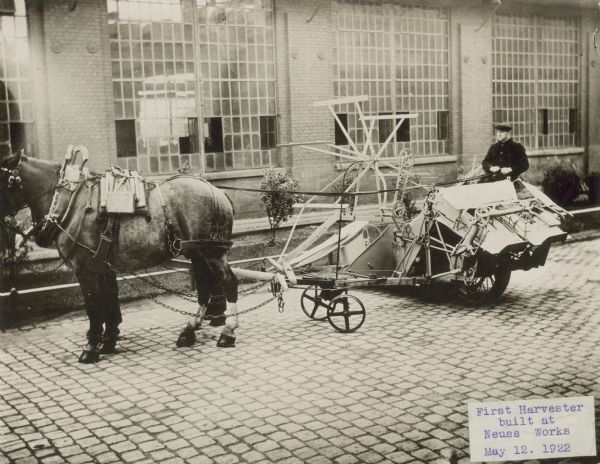 Left side view of a man sitting on horse-drawn harvester outdoors near a factory building.