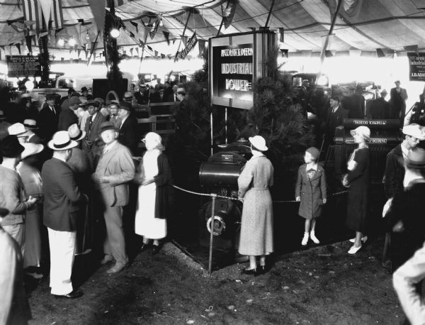 Slightly elevated view of a crowd of people gathered around engines and power units displayed beneath a tent at the Indiana State Fair. An electric sign flanked by two evergreen trees states: "McCormick-Deering Industrial Power."