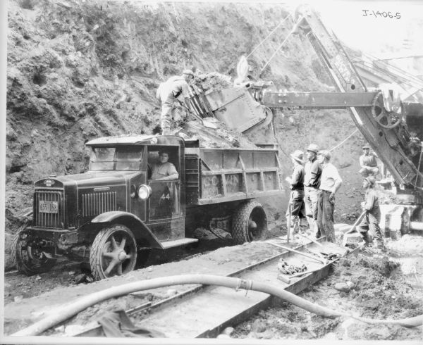Group of men working with a Bucyrus-Erie power shovel and dump truck. Hall-Scott powered dump truck with chain drive. 4 Cylinder, Overhead Cam, Hall-Scott engine. 3+2 speed transmission. The truck has a New York license plate, and on the side is painted: "Brooklyn, New York."
