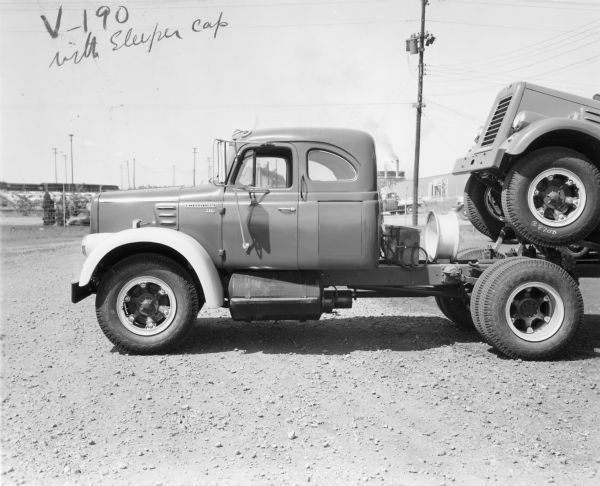 Driver's side view of a V-190 with sleeper cab parked in a lot. Spare wheel rim mounted on frame. The V-190 is pulling another V-190 to a dealer. Buildings are in the background.