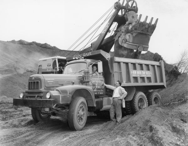 RDF 230 H tandem axle dump truck. Cummins diesel engine. 1200 x 24 tires. A man stands near the side of the driver's side door which has a sign reading: "Cinque's Trucking Company." The driver sits in the truck smoking a cigarette. A Bucyrus-Erie power shovel is dumping a load into the bed of the truck.