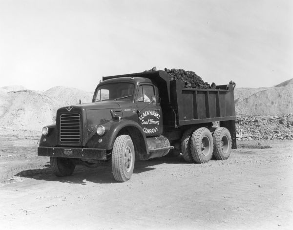 International VF-192 tandem dump truck with load of coal. Powered by V8-478 CID gas engine. Painted on the driver's side door: "Black Nugget Coal Mining Company." A driver sits in the driver's seat.