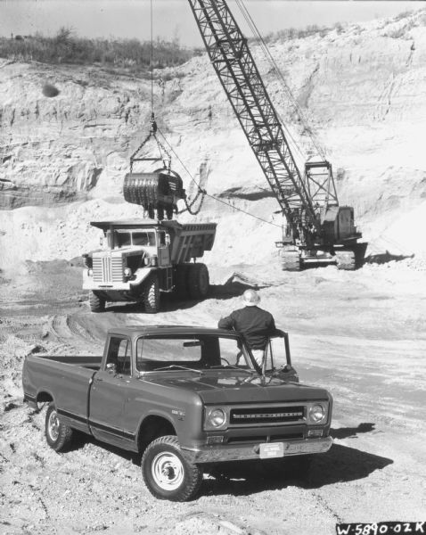 Slightly elevated view of a man standing with a 1/2 ton D-Line Model 1100-D 4x4 pickup. In the background a crane is loading an International Payhauler. Rock Truck.