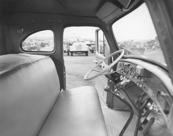 Interior view of R-Line sleeper cab from passenger side, driver's door open. Highway tractor at Fort Wayne factory yard and more vehicles are parked in the background.
