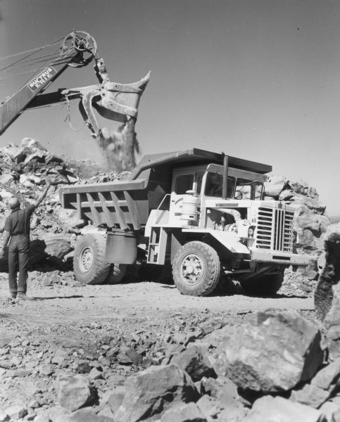 A man stands in the foreground on the left with arm raised while a Bucyrus-Erie power shovel dumps a load into a Model 65 Payhauler. Off road rock truck. The Model 65 was the smallest of the Payhaulers line.