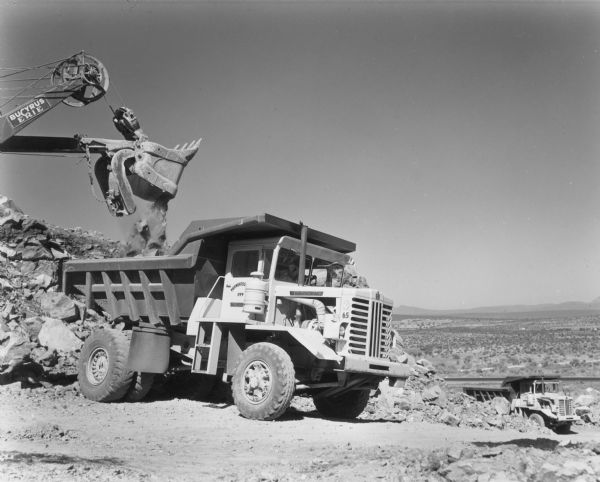 Right side view of a Model 65 Payhauler outdoors. A Bucyrus-Erie power shovel is dumping dirt and rock into the dump truck. The Model 65 is the smallest of the payhauler line. Another Payhauler is in the background, and beyond is a valley.