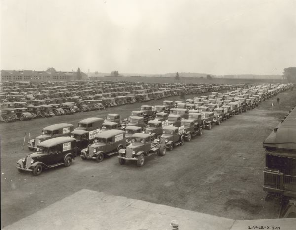 Elevated view of 153 C-Line Trucks parked in rows, ready for driveaway to the St. Louis branch. Total value of trucks was $142,880.91. The trucks have numbers on the passenger side of the windshield. Some 1/2 ton pickups are stacked on top of C-30 1 1/2 tons. There is a caboose of a railroad train parked on the right.