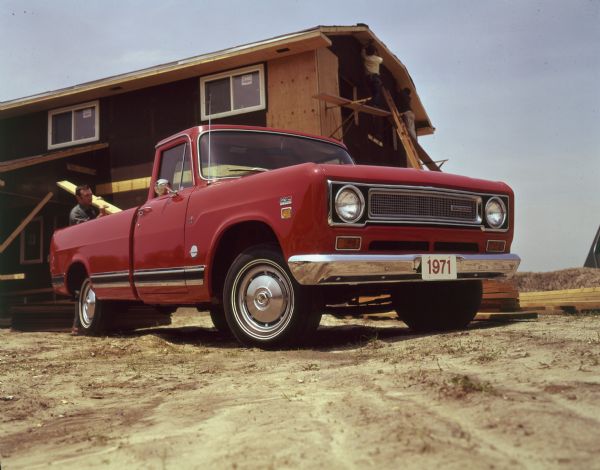 Three-quarter view from front of passenger side of 1110 Model half-ton pickup. Long wheel base, and 8 foot box. I-beam front axle. A man is holding a piece of lumber near the back of the pickup. Other men work on the construction of a building in the background.