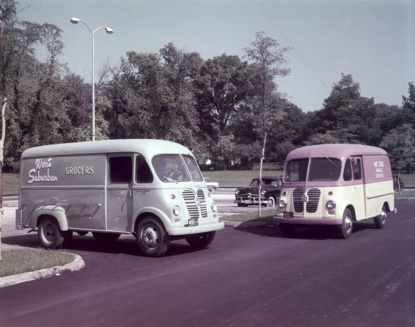 Two Metros in a parking lot. On the left, a man sits in the driver's seat of a S-150 Metro with a sign painted on the side reading: "West Suburban, Grocers." On the right a man is sitting in the driver's seat of a two-tone S-120 Metro with a sign on the side reading: "Metro Parcel Service."