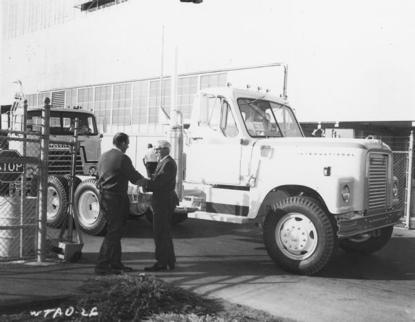 Right side view of International Truck passing through the gate leaving San Leandro plant. Two men shaking hands are on the left.