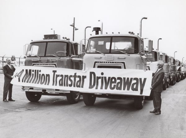 Cab overs leaving San Leandro factory. Two men hold a banner in front of the two rows of trucks that reads: "$4 Million Transtar Driveaway."