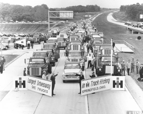 Elevated view of the opening of the driveaway on Interstate 70. Men stand holding signs that read: "Largest Driveaway in IH Truck History, 946 Trucks, Springfield, Ohio."