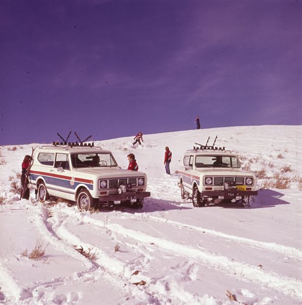 Scout II vehicles parked in the snow. Decals on the side read: "US SKI TEAM." Men and women in ski clothing stand nearby. One woman is skiing down the hill in the background.