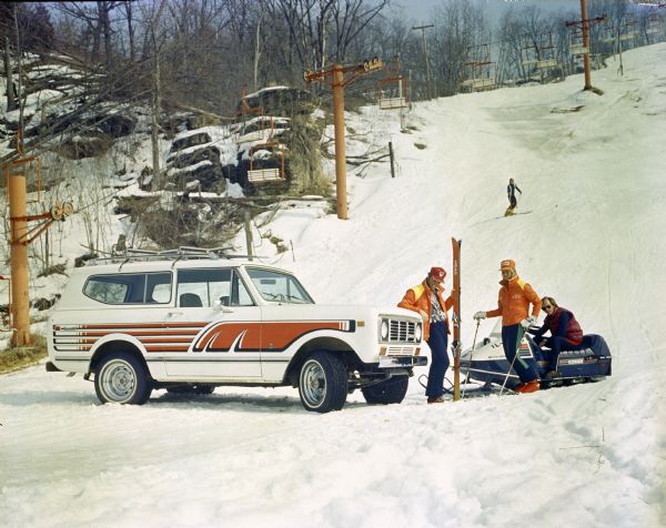 View up ski hill towards people standing near a Scout II in the snow. One man sits on a Quiet Flite Evinrude snowmobile near a man holding a pair of Head snow skis and a woman standing on skis. In the background another man is skiing down the hill.