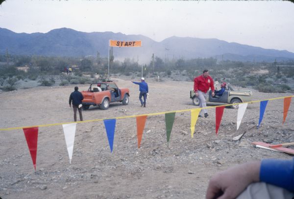 View over flag banner towards men in a dirt area near a "START" sign with International vehicles. Some of the men riding in the trucks are wearing helmets. Mountains are in the background.