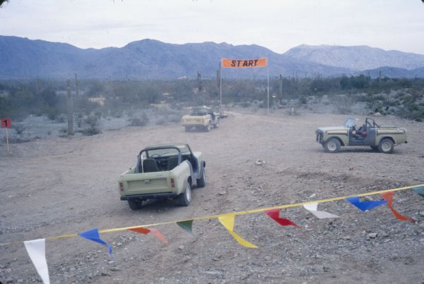 Slightly elevated view over flag banner of International vehicles in a dirt area near a "START" sign near a road. In the distance are mountains.