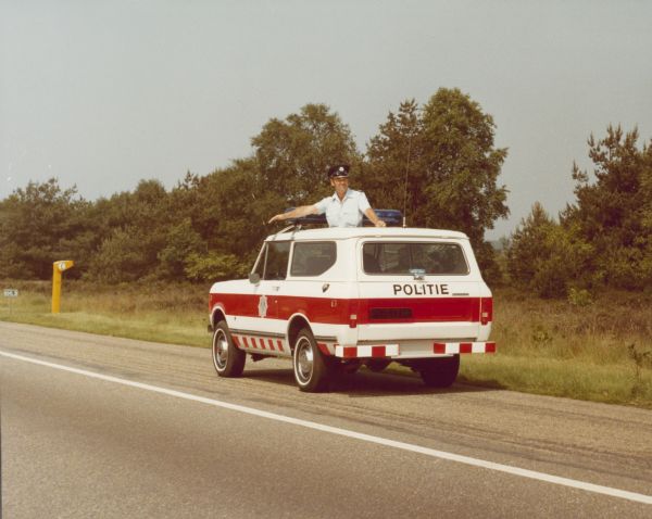 Three-quarter view from rear of diver's side of Police Unit parked on the side of a road. A man wearing a uniform is standing on the front seat through the open sunroof of the truck.