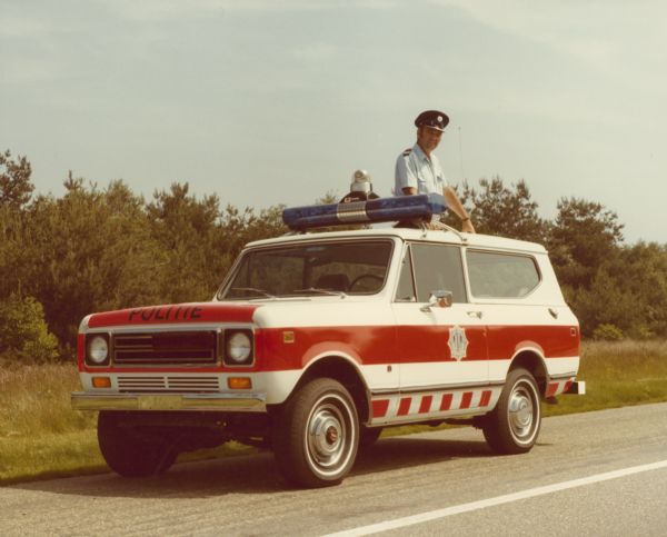 Three-quarter view from front of diver's side of Police Unit parked on the side of a road. A man wearing a uniform is standing on the front seat through the open sunroof of the truck.