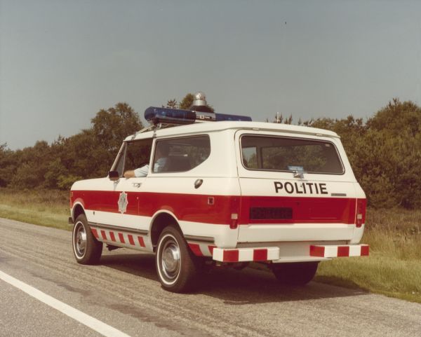Three-quarter view from rear of diver's side of Police Unit parked on the side of a road. A man wearing a uniform is sitting in the driver's seat.