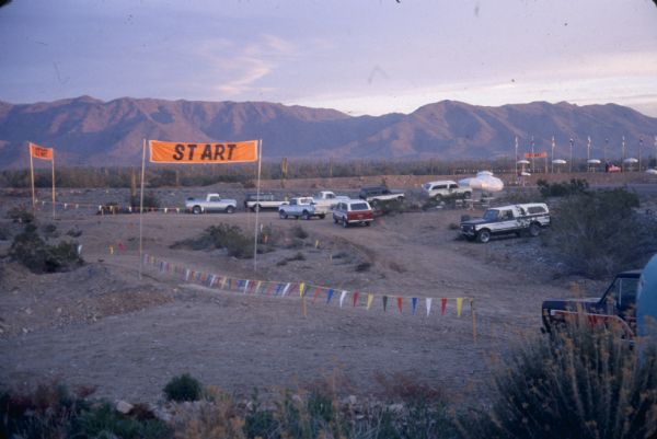 View across dirt parking area towards men with International vehicles. There are "START" signs on poles near a dirt road. Tables with umbrellas, and flags on flagpoles are on the far right. Mountains are in the distance.