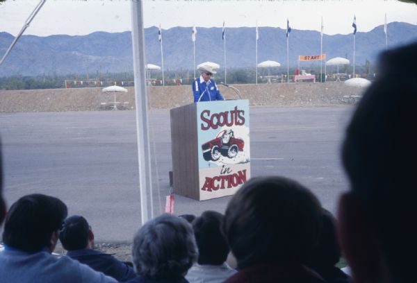 View from audience looking towards a man wearing a cowboy hat. He is standing at a podium in a parking lot. The sign on the podium reads: "Scouts in Action." In the background are tables with umbrellas, flags on flag poles, and a "START" sign. Mountains are in the distance.