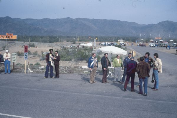 Slightly elevated view of groups of men standing outdoors. Some of the men are standing along a paved road in the foreground. In the background on the left is a "START" sign. International vehicles are parked in the background. Mountains are in the far background.