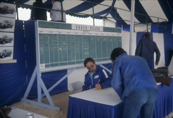 Men in tent working at a table. Photographs of Scouts are on the left. A chalkboard titled "ORDER BOARD" lists the "Northwest Region." Behind the sign is a partition against a grandstand, where two people sit looking out.