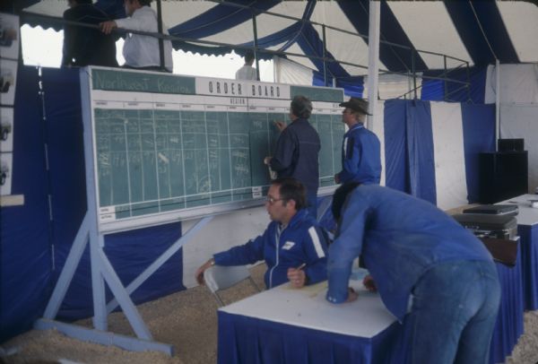 Men in tent working at a table. Photographs of Scouts are on the left. A chalkboard lists the "Northwest Region." Behind the sign is a partition against a grandstand, where two people sit looking out.

