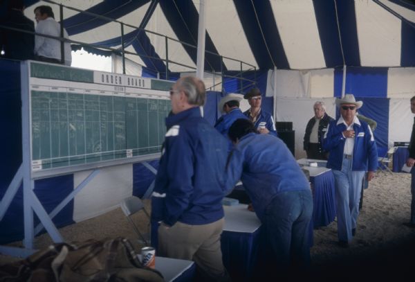 Men in tent working at a table. Photographs of Scouts are on the left. A chalkboard lists the "Northwest Region." Behind the sign is a partition against a grandstand, where two people sit looking out.