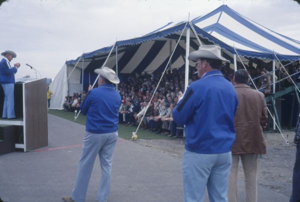 Men stand near a tent. In the background on the left a man stands at a podium addressing an audience sitting in the tent.