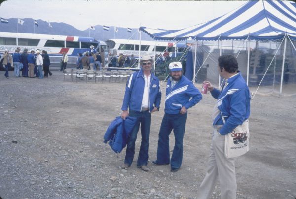 Three men stand in the foreground. One of the men wears a "Scout" cap. Chairs are set up in rows near a podium and a tent. Behind the podium people are gathered near Greyhound buses. Flags fly on flagpoles, including an "IH" flag. In the far background are mountains.