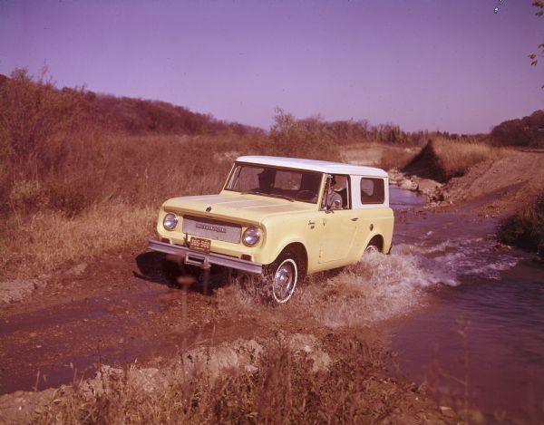 Three-quarter view from front towards a man driving a Scout on a road crossing a stream. Another man is sitting in the passenger seat.
