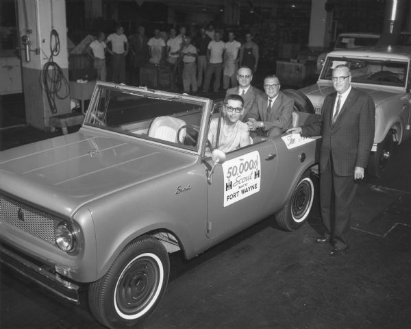 Wayne Van Ryn, final assembly employee, drives the rear-wheel drive model with 4-passenger capacity and bucket front seats off the line. Two men, V.A. Hart and M.E. Hayhurst, sit in the back seat. Standing next to the Scout is H.A. Torgersen. A group of factory workers is standing in the background.