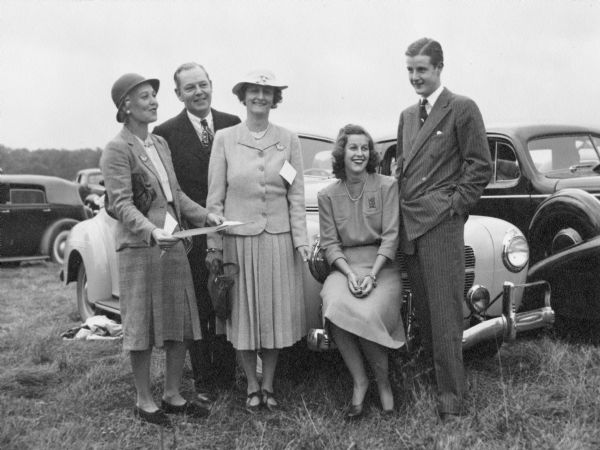 Outdoor group portrait of the McCormick Family. Original caption on the back reads: "Lake Forest 1941 or 1942. Aunt Lucy, Mr. & Mrs. McCormick, Hope and Brooks." The family is posing standing and sitting in front of a car in an open field surrounded by other cars.